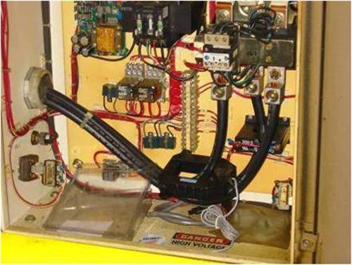 Interior of an electrical control panel with wiring, relays, and a current transformer clamped around supply cables, marked with a high voltage warning label at the bottom.